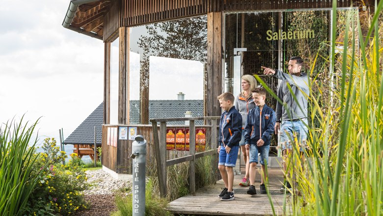 Part of the "Water of Life" theme trail, © Erlebnisalm Mönichkirchen, Martin Fülöp Family on a wooden path in front of a building with the inscription 'Salaerium'.