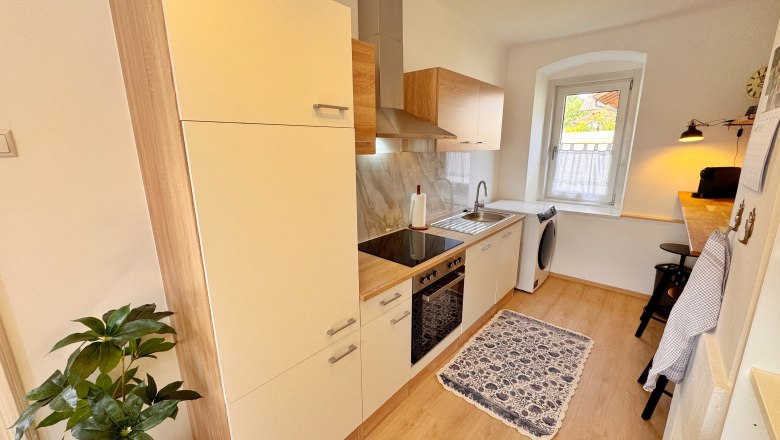 Kitchen, © Christoph Menhofer Modern kitchen with wooden and white units, stove, sink, washing machine and window. Plants and carpet as decoration.