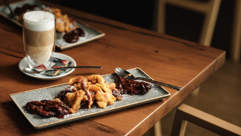 W4, © Weinviertel Tourismus / Michael Reidinger A plate of Kaiserschmarrn and stewed plums on a wooden table with a glass of latte macchiato next to it.