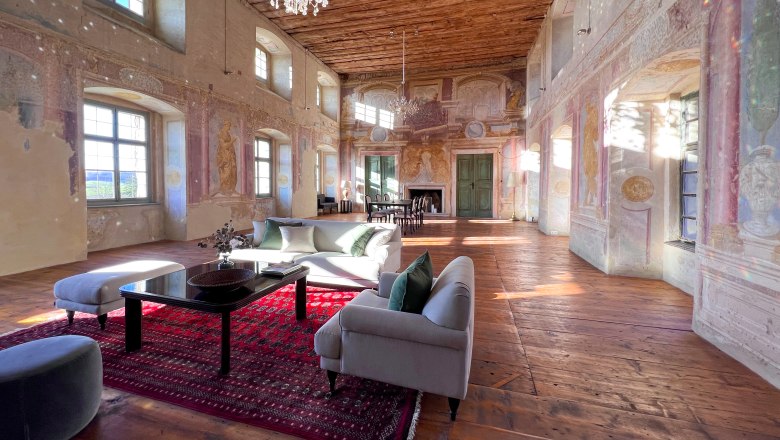 Ballroom, © Schloss Haggenberg A historic ballroom with a wooden floor, painted walls and a chandelier. Modern furniture and a red carpet are placed in the room.
