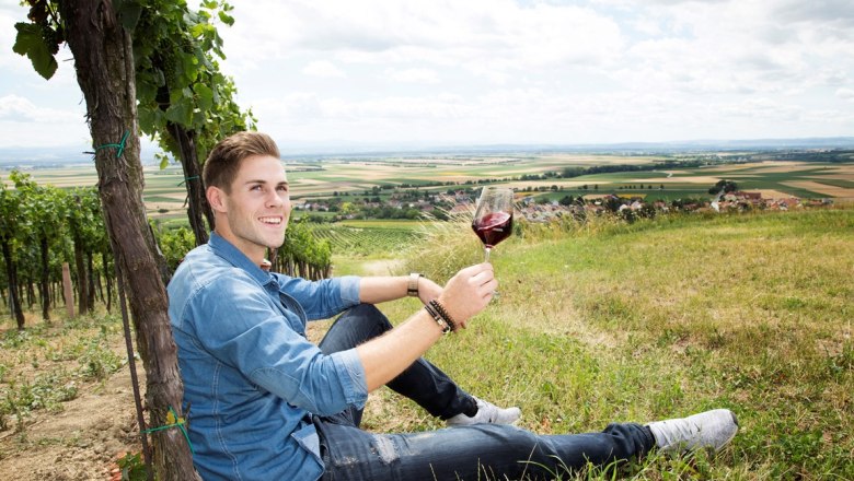 Patrick Kerner, © Weingut Kerner A man sits in a vineyard holding a glass of red wine. A vast landscape can be seen in the background.