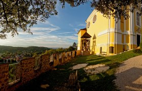 Viewpoint at the mountain church in Pitten, © Wiener Alpen, Franz Zwickl Yellow church on a hill with a view of a village and wooded hills in the background.