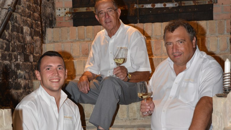 Waldschütz family, © Familie Waldschütz Three men in white shirts sit on a stone staircase holding wine glasses.