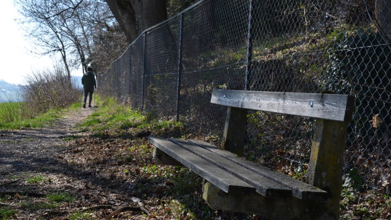 hiking-path-artstetten-copy, © Karl Temper A hiking trail with a wooden bench and a chain-link fence. A person is walking in the distance.