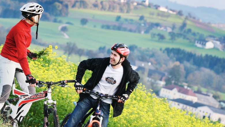 Cyclist near Euratsfeld, © weinfranz.at Two cyclists with helmets are standing in a flowering field with a green landscape in the background.