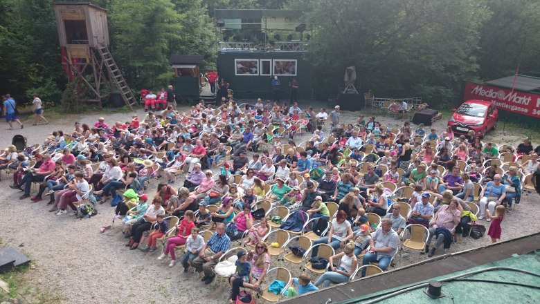 Quarry auditorium, © Theater Purkersdorf An audience sits outside on chairs in a quarry, surrounded by trees and a red car with Media Markt advertising.