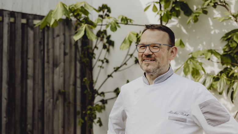 Landlord Markus Bsteh, © Niederösterreich Werbung/Sophie Menegaldo A man in chef's clothes stands smiling in front of a wooden wall with plants.