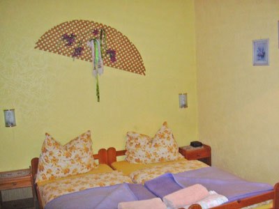 Room view, © Jeschko A cozy bedroom with yellow walls, a double bed with patterned pillows and a decorative wall fan.