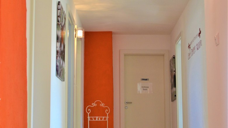 staircase, © Gluschitz-Goebel A hallway with a wooden floor, white walls and an orange wall. There is a sign on the wall with the words 'Caution step'.