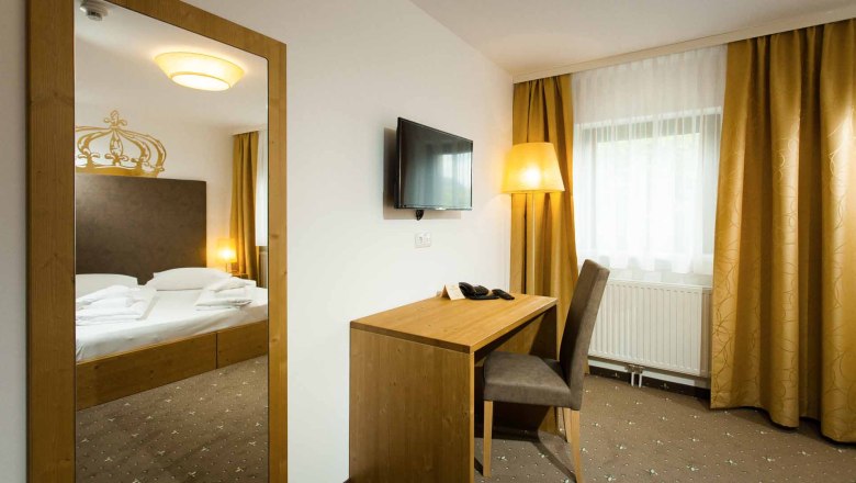 Double room, © Hotel Kaiser Franz Josef A hotel room with a double bed, a desk and chair, a TV on the wall and yellow curtains.