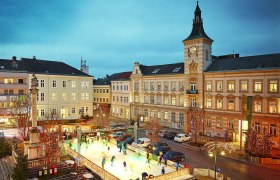 Advent magic at the ice rink, © Alexander Bernold Ice rink in a town at dusk, surrounded by historic buildings and Christmas lights.