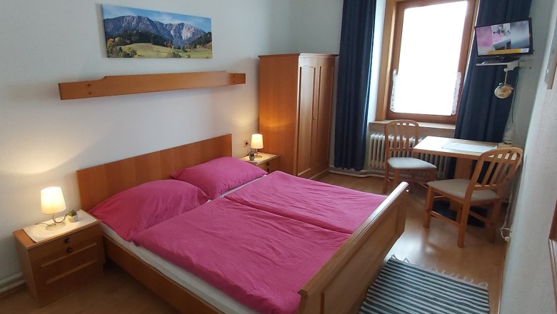 Double room, © Martin Zwinz A cozy double room with wooden furniture, a double bed with pink bed linen, a table with two chairs and a small TV on the wall.