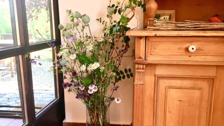 Natural garden exit, © Fam. Ritz A bunch of wild flowers in a vase next to a piece of wooden furniture, with a view of a garden through a window.