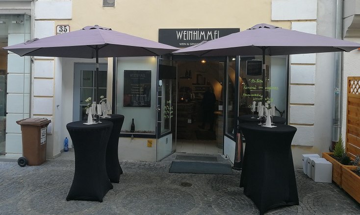 The place close to the sky, © Roman Zöchlinger Entrance to a wine bar with two bar tables and parasols in front of it.