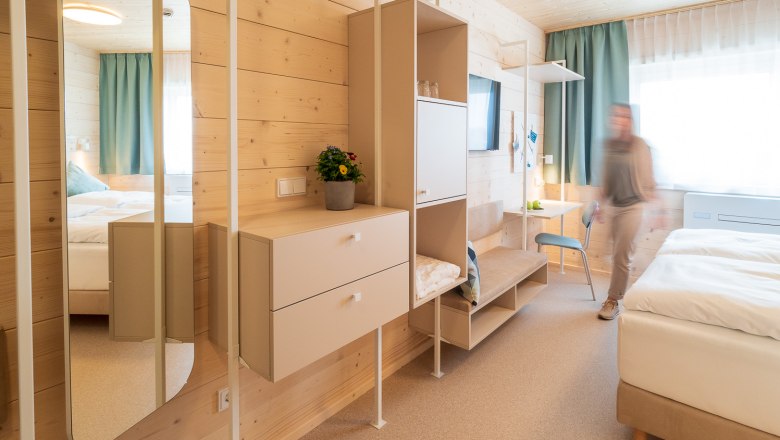 Sufficient storage space, © freilich / IsaBella Fritsche Modern hotel room with wooden paneling, mirror, wardrobes and a bed. One person moves around the room.