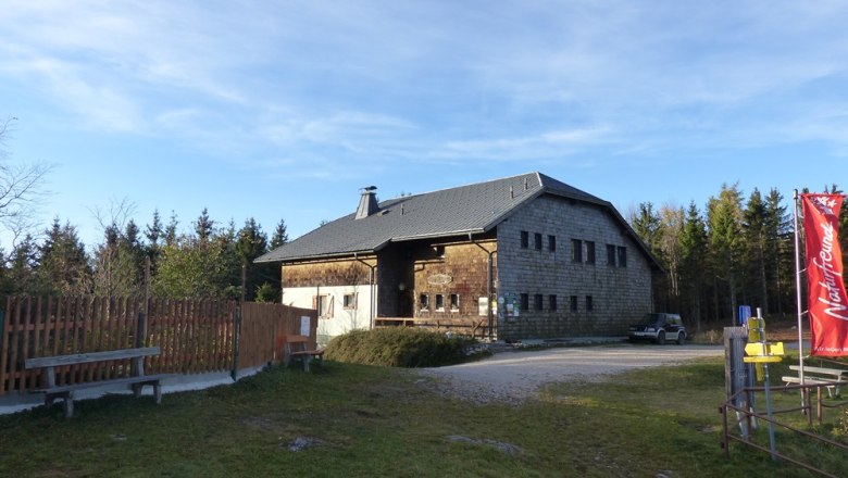 Öhler shelter, © Mario Putz A rustic building with wooden walls and a gray roof, surrounded by trees. A red flag is on the right of the picture.