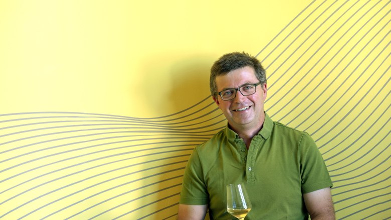 Thomas Buchmayer, © Michael Himml Man with glasses and green shirt holds a wine glass in front of a yellow background with a line pattern.