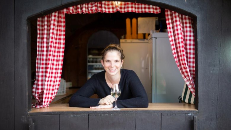 Boss and former wine queen, © Alexander Pfeffel Woman leaning out of a window with red and white checkered curtains, holding a glass of wine.