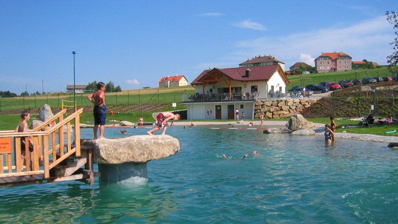 Nöchling bathing pond, © Nöchling Children playing at a swimming pond with a jetty and rocks, a building and cars in the background.