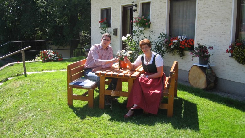 A warm welcome, © Wachahof Two people are sitting at a wooden table in the garden in front of a house with flowers.