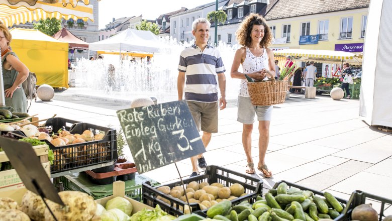 Tulln Naschmarkt, © Stadtgemeinde Tulln, Robert Herbst A couple walks through a market with vegetables and signs. Market stalls and a fountain can be seen in the background.
