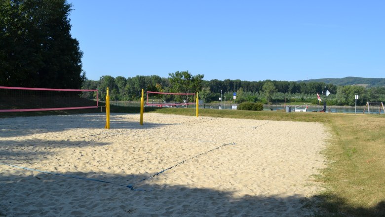 TD beach volleyball court, © Stadtgemeinde Korneuburg An empty beach volleyball court with yellow posts and a pink net, surrounded by trees and a river in the background.