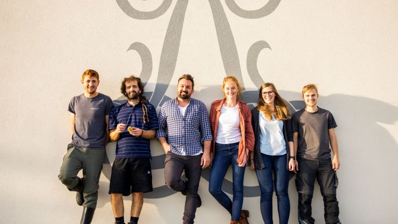Team, © Weingut Liechtenstein Six people stand smiling in front of a wall with a decorative pattern.