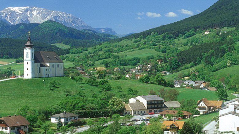 The Landgasthof Jautschnig at the Maria Kirchbüchl pilgrimage church, © Gasthaus Jautschnig Landscape with a church on a hill, surrounded by green fields and mountains in the background.