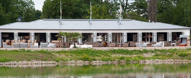 Terrace view, © Diamond Club Atzenbrugg A long building with patio furniture in front of a pond, surrounded by trees.