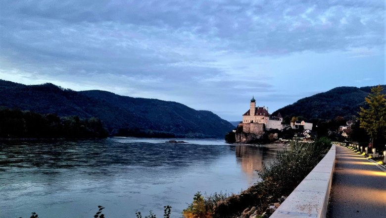 Treppelweg in the direction of the castle, © Szilágyi An illuminated path leads along a river to a castle on a hill surrounded by wooded mountains.