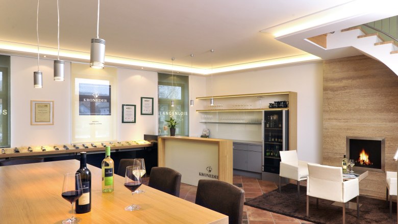 Wine & Living Kroneder, © Familie Kroneder Interior view of a modern wine tasting room with wooden table, wine glasses, fireplace and shelves.