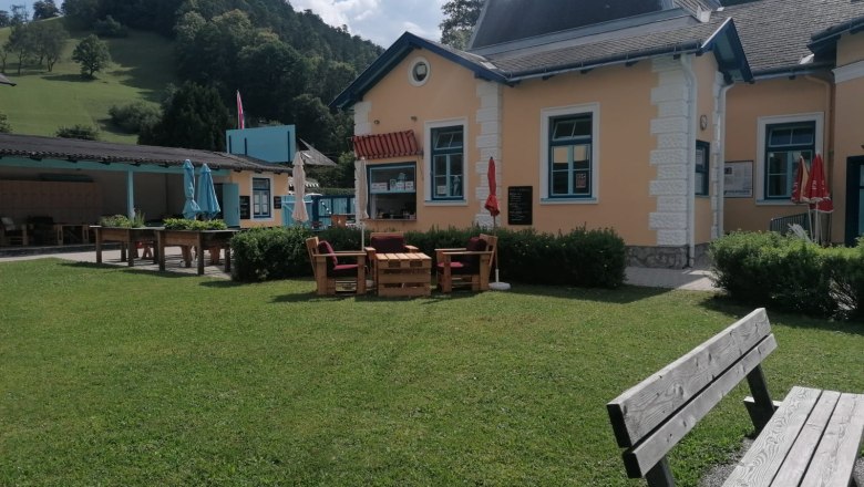 photo-bathroom-2, © Gemeinde Gutenstein A yellow building with blue window frames, surrounded by a green meadow and benches, in front of a wooded hill.