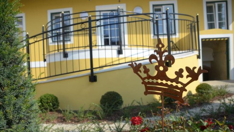 Garden, © Julia Pfeiffer / Fam. Engelbrecht A yellow building with a balcony and a garden with red roses and a metal crown as decoration.