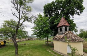 Sacred stone, © Weinstraße Weinviertel Small chapel with a red tiled roof in a green landscape, surrounded by trees and benches.