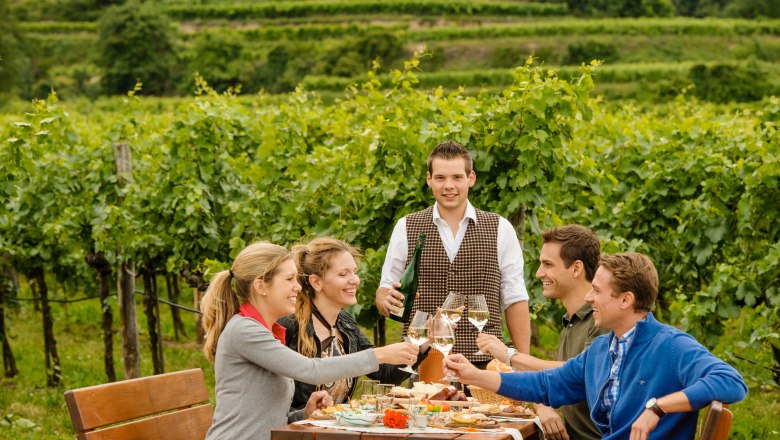 Heurigen catering, © Rita Newman A group of people are sitting at a table in the vineyard, clinking glasses of wine. A waiter stands next to them with a bottle of wine.