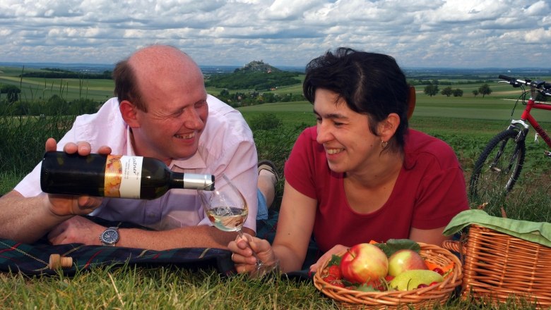 Doris and Reinhard Rieder, © Seymannfilm A couple having a picnic in a meadow with wine and a fruit basket, in the background a hilly landscape and a bicycle.