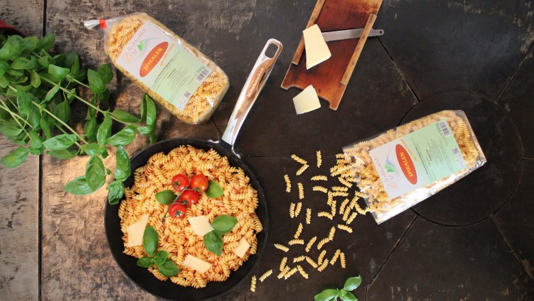 Pasta at its finest, © Familie List A pan of spiral noodles, tomatoes and basil on a wooden table, surrounded by pasta wrappers and a cheese slicer.