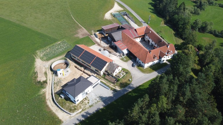 Knappenhof Hahn - Aerial view, © Familie Hahn Aerial view of a farm with several buildings and solar panels on a roof, surrounded by fields and forests.