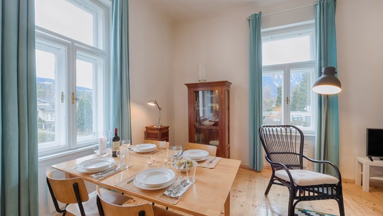 Top 5 - Living room, © VP FeWo OG Dining area in a living room with wooden table, chairs, display cabinet and large windows with blue curtains.