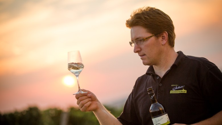 Winemaker Wolfgang Müllner, © Astrid Bartl A man holds a wine glass and a wine bottle in the vineyard at sunset.