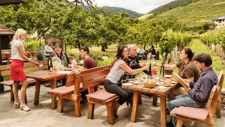 Outside at the Strawanzer wine tavern, © Rita Newman People sit at wooden tables outside, surrounded by vineyards and trees, while a waitress serves drinks.