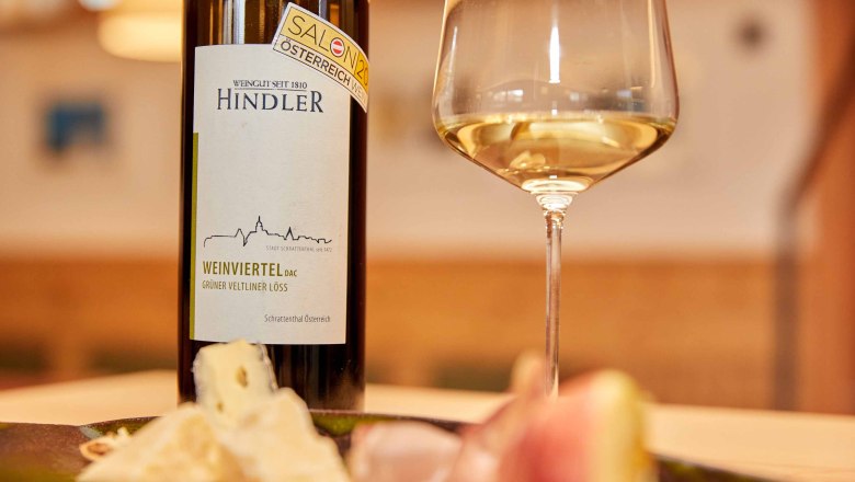Hindler Winery, © Weingut Hindler Wine bottle and glass with white wine, cheese and ham in the foreground.