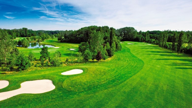 Golf Club Gutenhof, © Golf Club Gutenhof Green golf course with bunkers and pond, surrounded by trees under a blue sky.