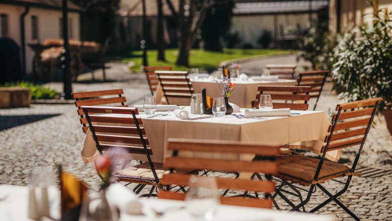 Guest garden in the inner courtyard, © Niederösterreich Werbung / Daniela Führer A sunny guest garden with wooden tables and chairs, set with white tablecloths and glasses, in a paved courtyard.