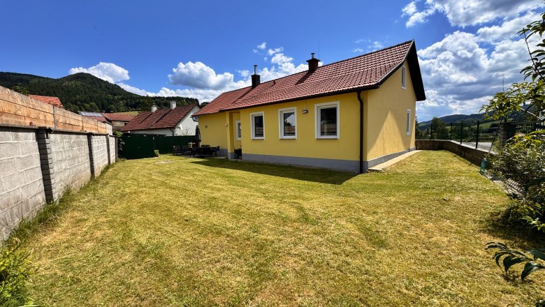 Garden, © Christoph Menhofer A yellow house with a red roof in a garden with a lawn, surrounded by a wall and mountains in the background.