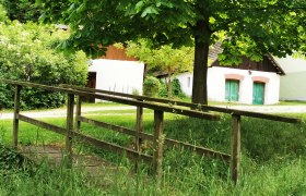 Romantic wine cellar lane in the middle of the Schmidatal valley, © Weinstraße Weinviertel A small wooden bridge leads across a meadow to white buildings with green doors, surrounded by trees and bushes.