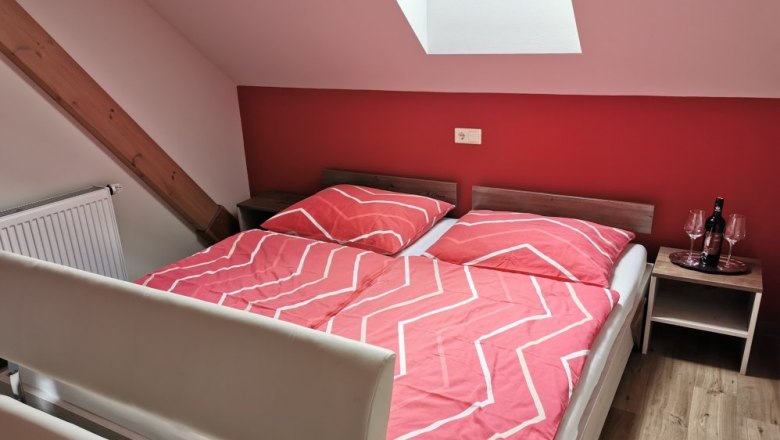 Vacation apartment "Zweigelt", © Weinhof Brauner Bedroom with red wall, double bed with red bed linen, skylight and bedside table with wine bottle and glasses.