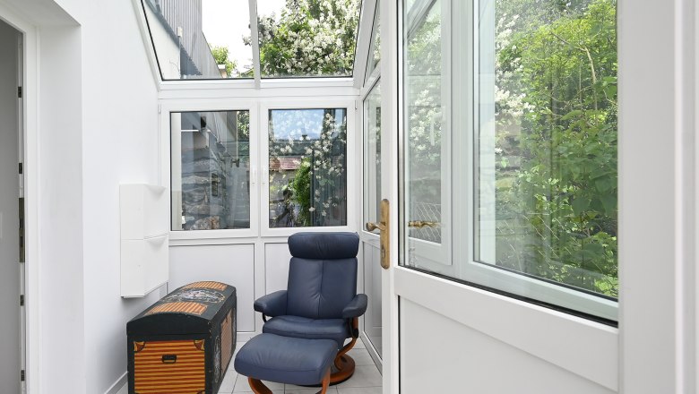 Anteroom, © Christine Kuppelwieser A bright anteroom with a blue armchair and a decorative chest, surrounded by large windows with a view of green plants.