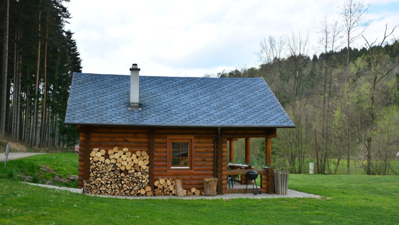 Log cabin - Gaul, © Karin Spiesmaier A log cabin with woodpile and barbecue in the countryside, surrounded by trees.