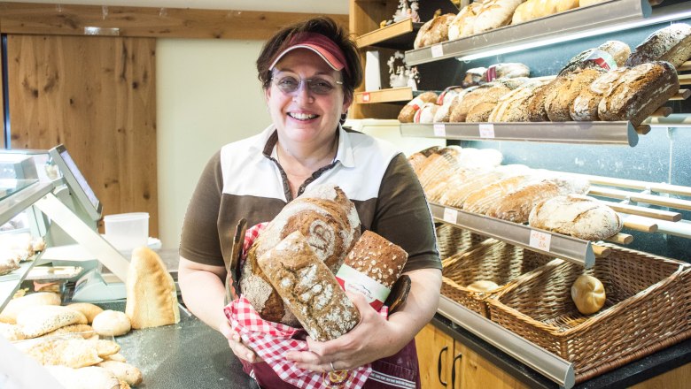Martina Birnbauer, © Clemens Trenker, BA A woman in a bakery holds several loaves of bread in her hands and smiles at the camera.